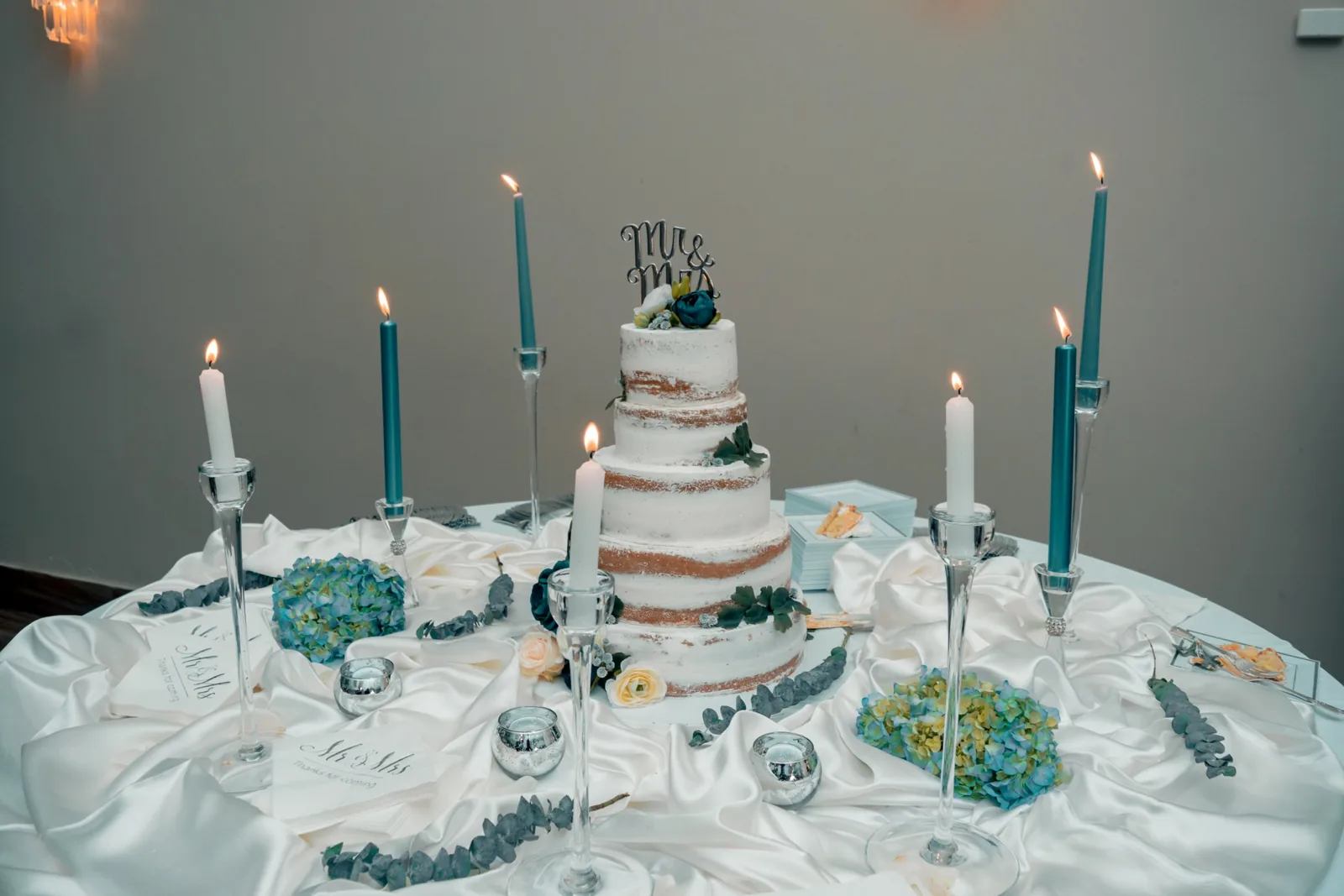 Three-tier semi-naked wedding cake with blue flowers and teal candles