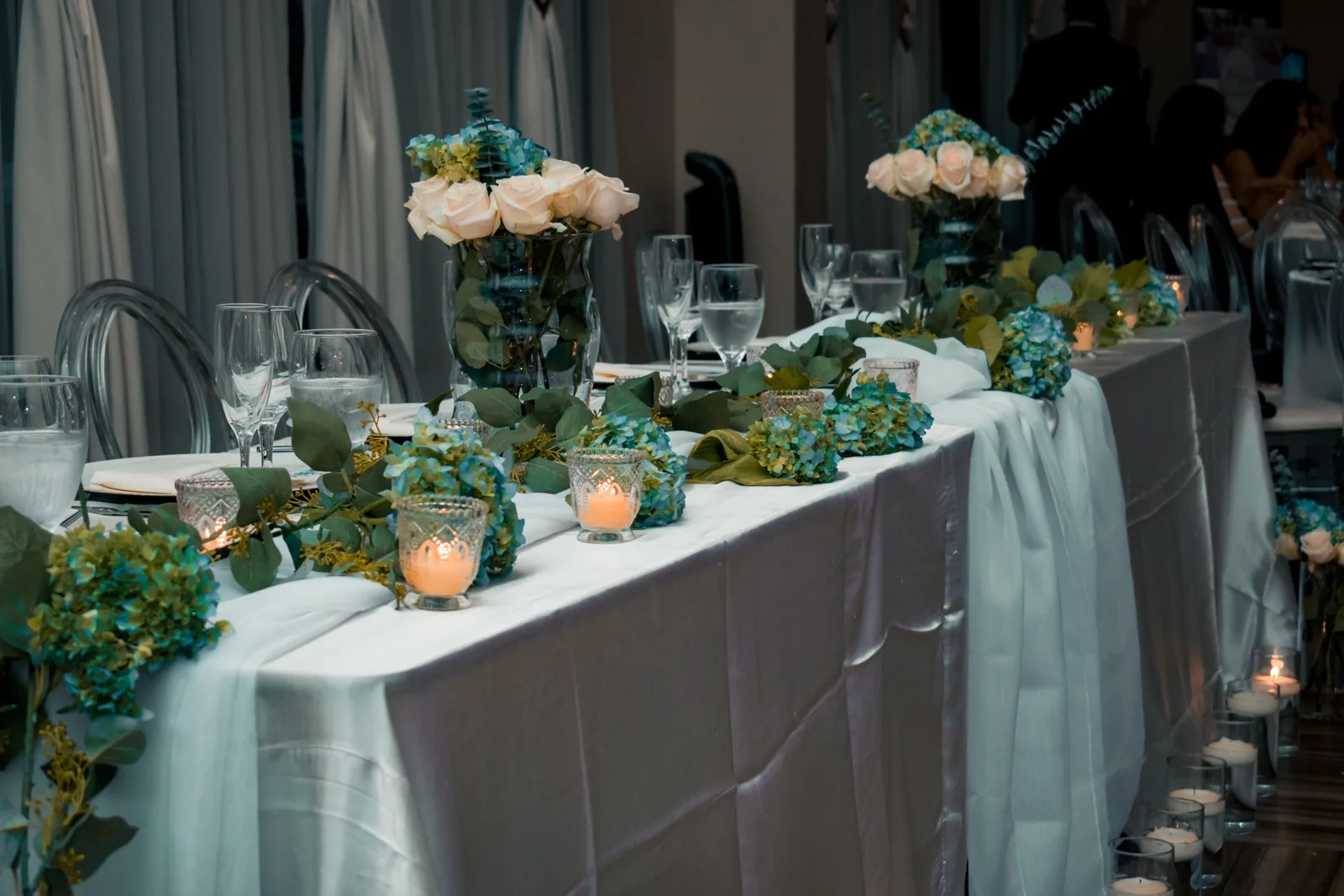 Head table set with glassware, white roses, hydrangeas and candles along a draped table