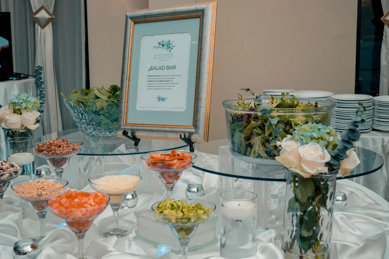 Salad bar with toppings in glass cups, glass bowls of greens and a framed menu sign