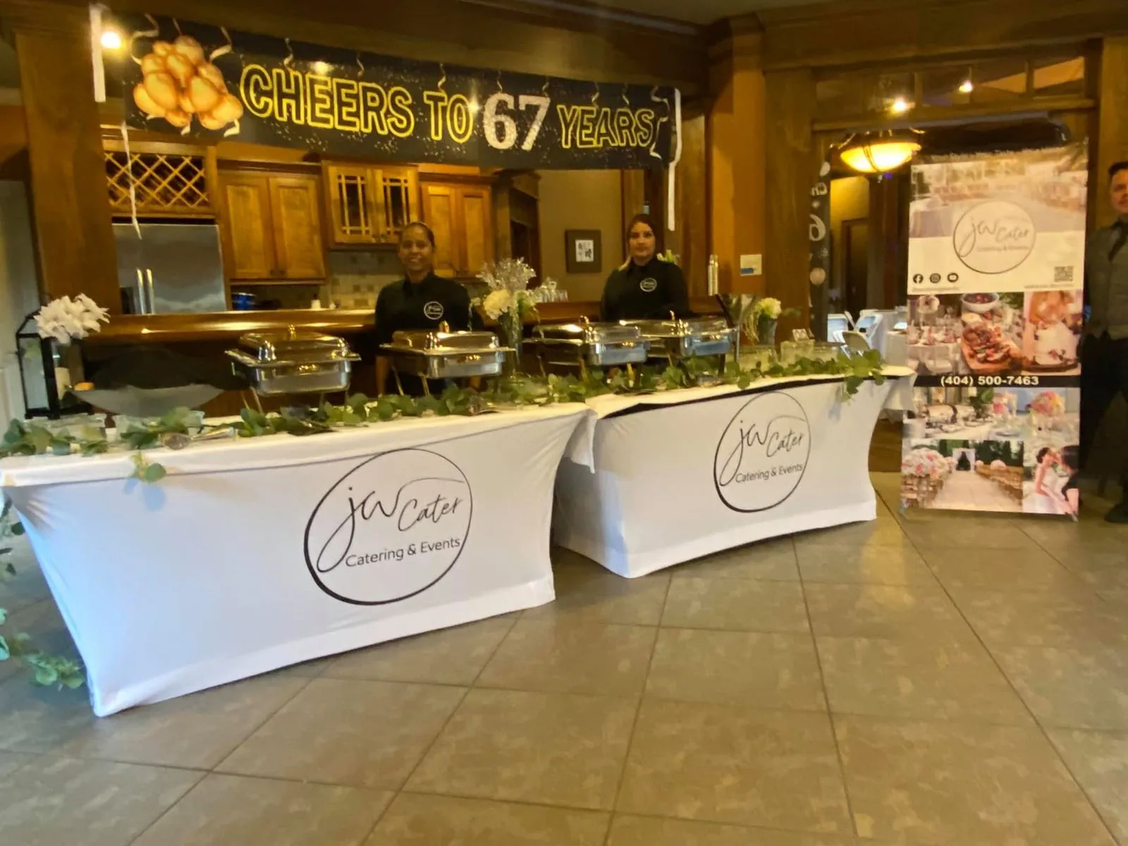 Buffet under a Cheers to 67 Years banner with two JW Cater staff