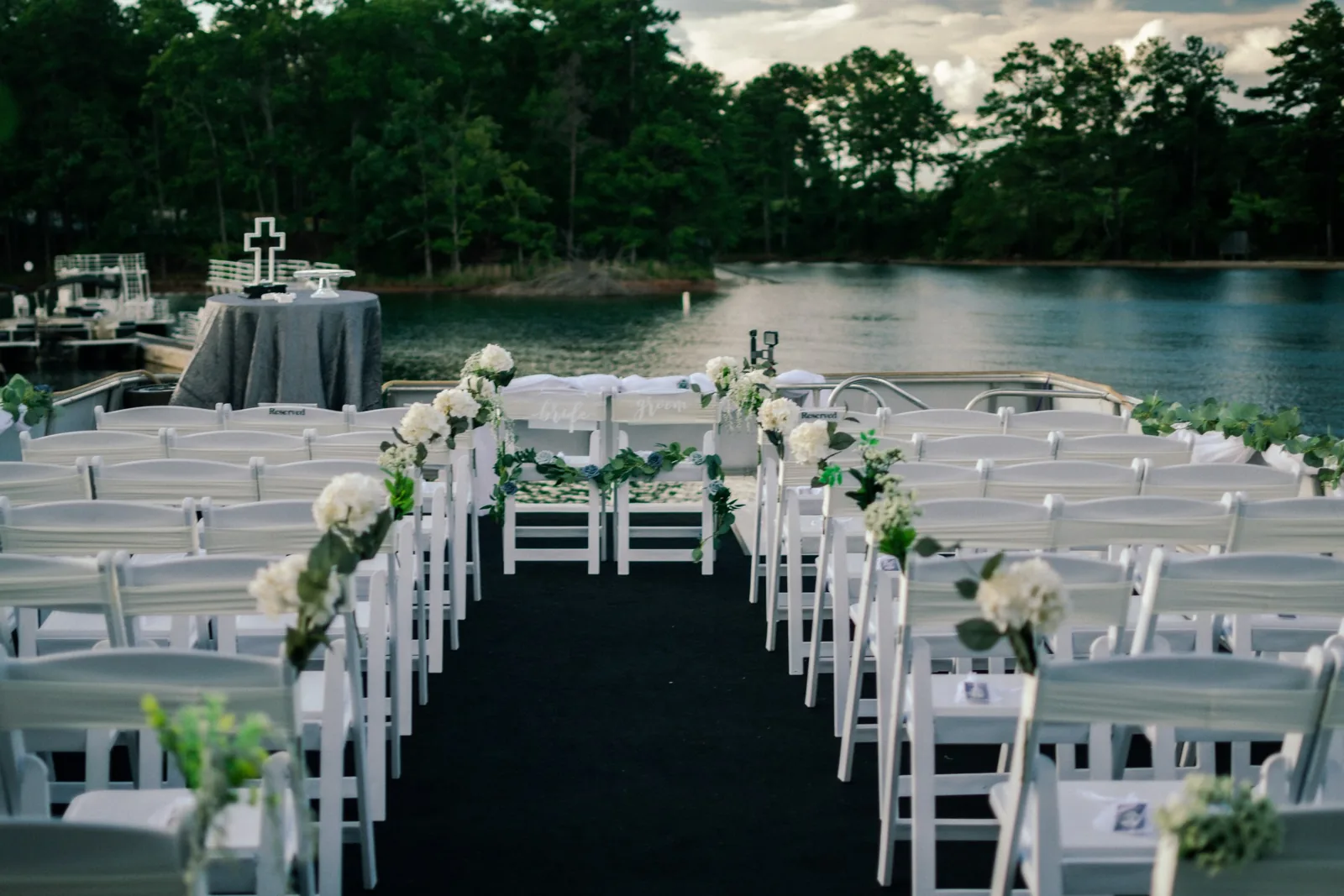 Lakeside wedding ceremony with white chairs and a greenery aisle on a dock