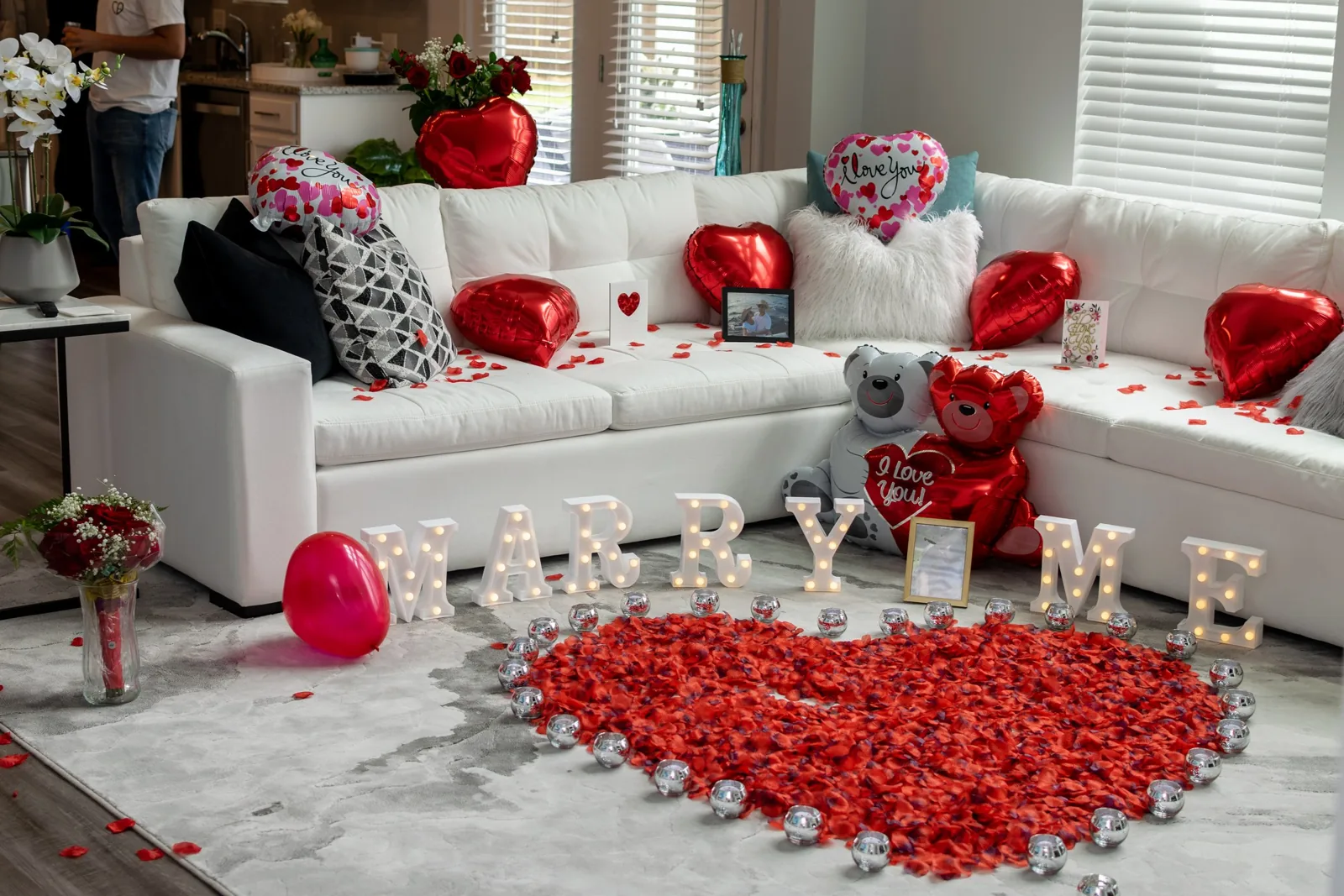 Marry me proposal setup with rose petals, red heart balloons and light-up letters